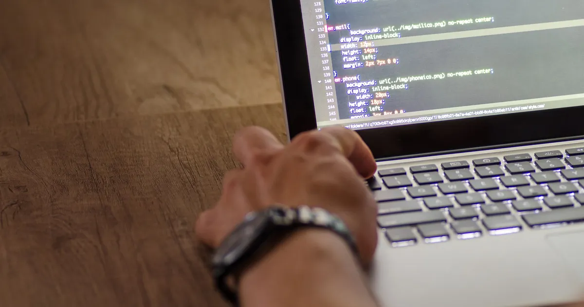 A developer's hand interacting with code on a laptop screen in a workspace setting.
