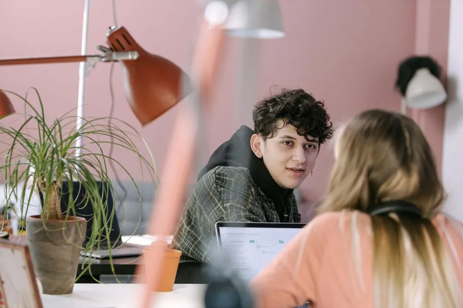 Two young professionals in a modern office space, discussing work over laptops and plants.